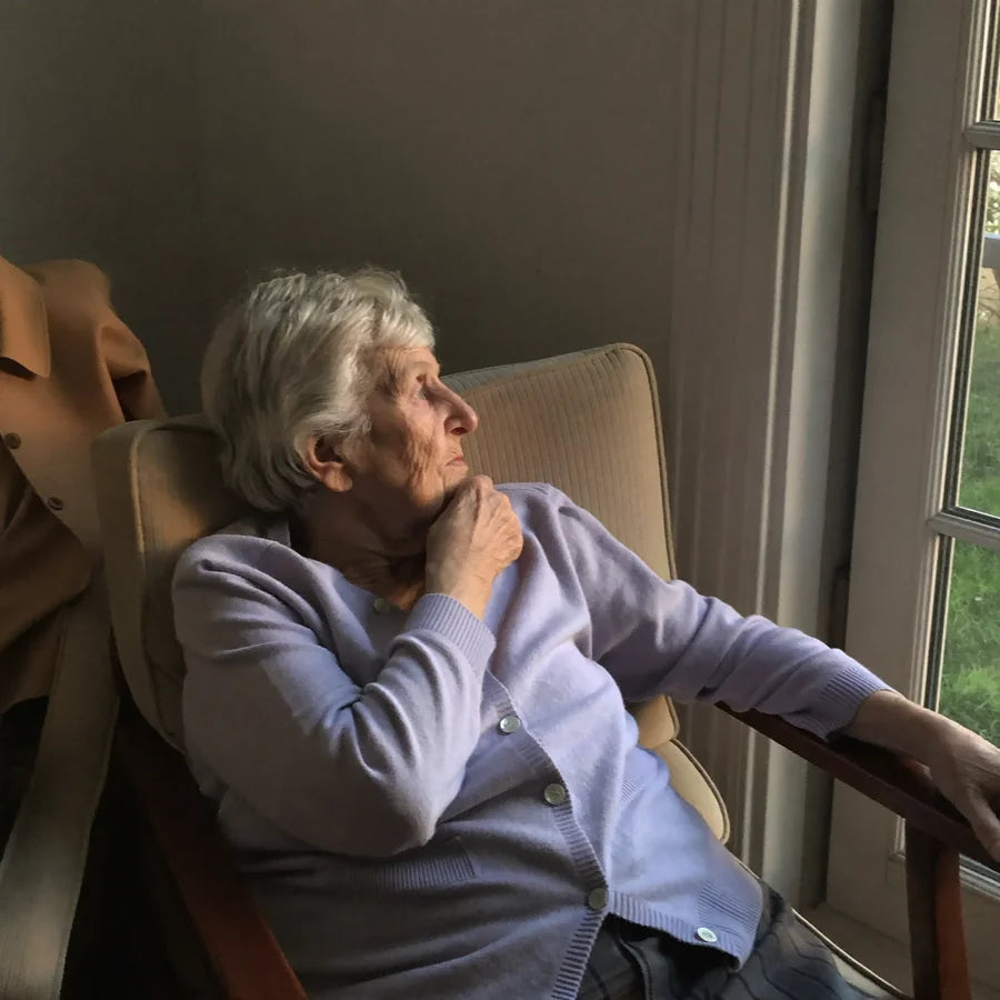 Une femme âgée assise seule dans son fauteuil, manteau et sac posés à côté, sans sortir