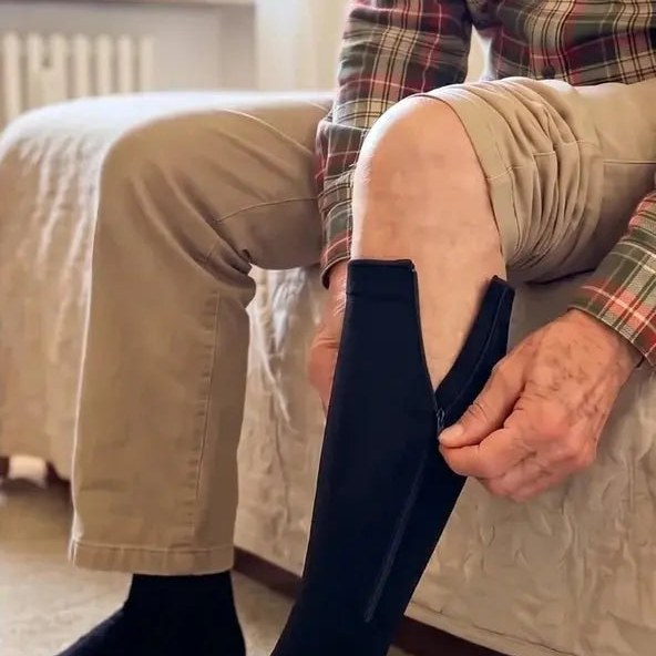 Assis au bord du lit, un homme âgé remonte seul la fermeture de sa chaussette Liberté noire.